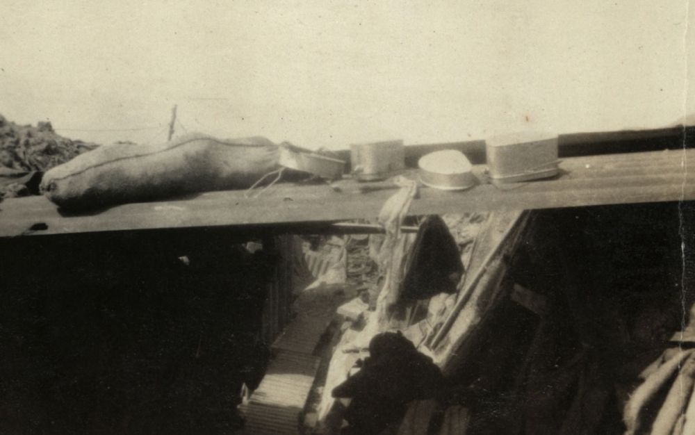 A trench in Armentières. A sandbag, mess tins and drinking panniers sit atop the corrugated iron while a soldier sleeps below. 1916.